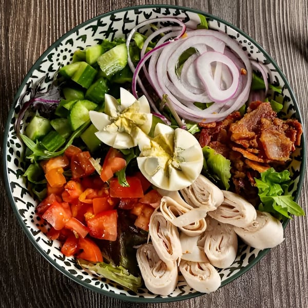 Overhead view of a salad with diced tomatoes, cucumbers, red onion, bacon, hard-boiled eggs, and rolled turkey.