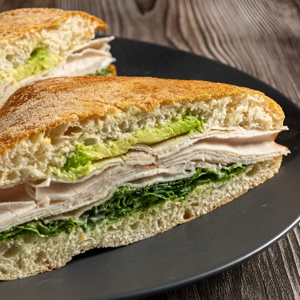A sliced turkey and avocado sandwich on ciabatta bread with lettuce, served on a dark plate.